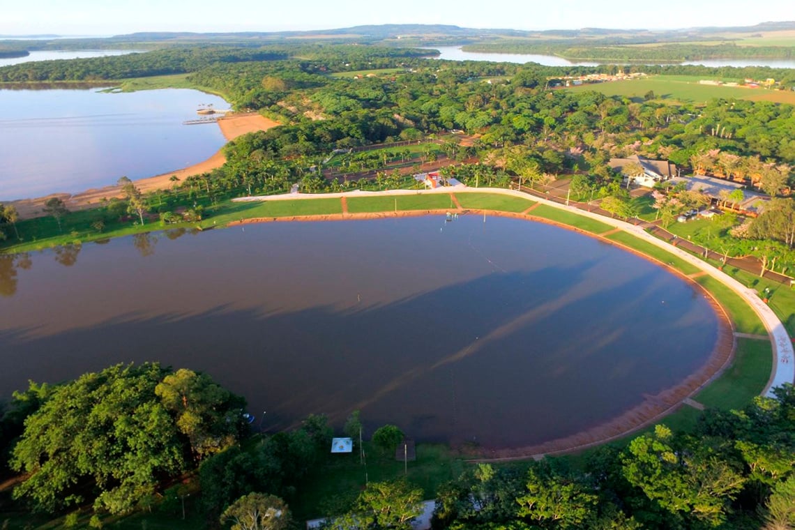 Balneário de Santa Helena no Lago de Itaipu ao entardecer: praia artificial com faixa de areia, água azul-esverdeada e árvores ao fundo
