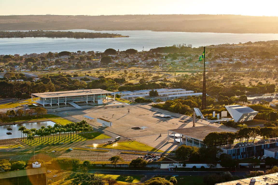 Vista panorâmica de Brasília ao pôr do sol, com áreas verdes, Lago Paranoá e o Congresso Nacional ao fundo.