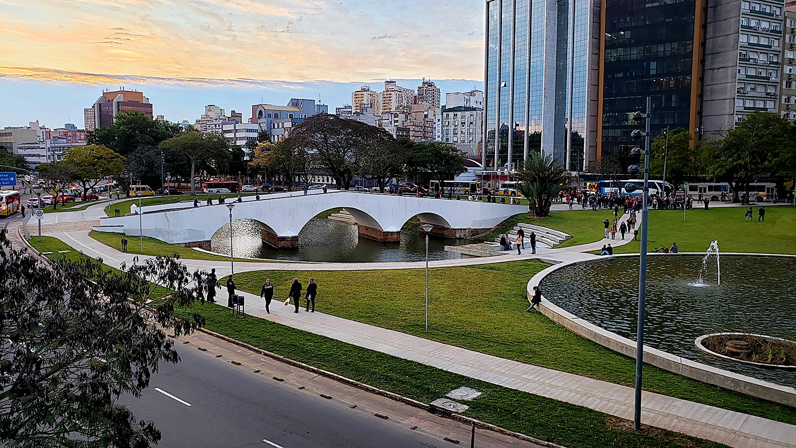 Paisagem urbana típica do interior gaúcho, com praça arborizada, lago e ponte branca ao fundo