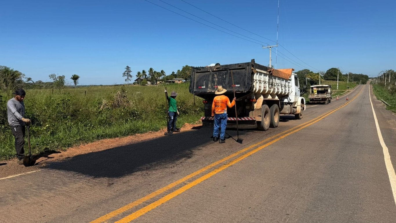 Equipe da Secretaria de Obras trabalhando em via urbana, com caminhão caçamba e rolo compactador ao fundo