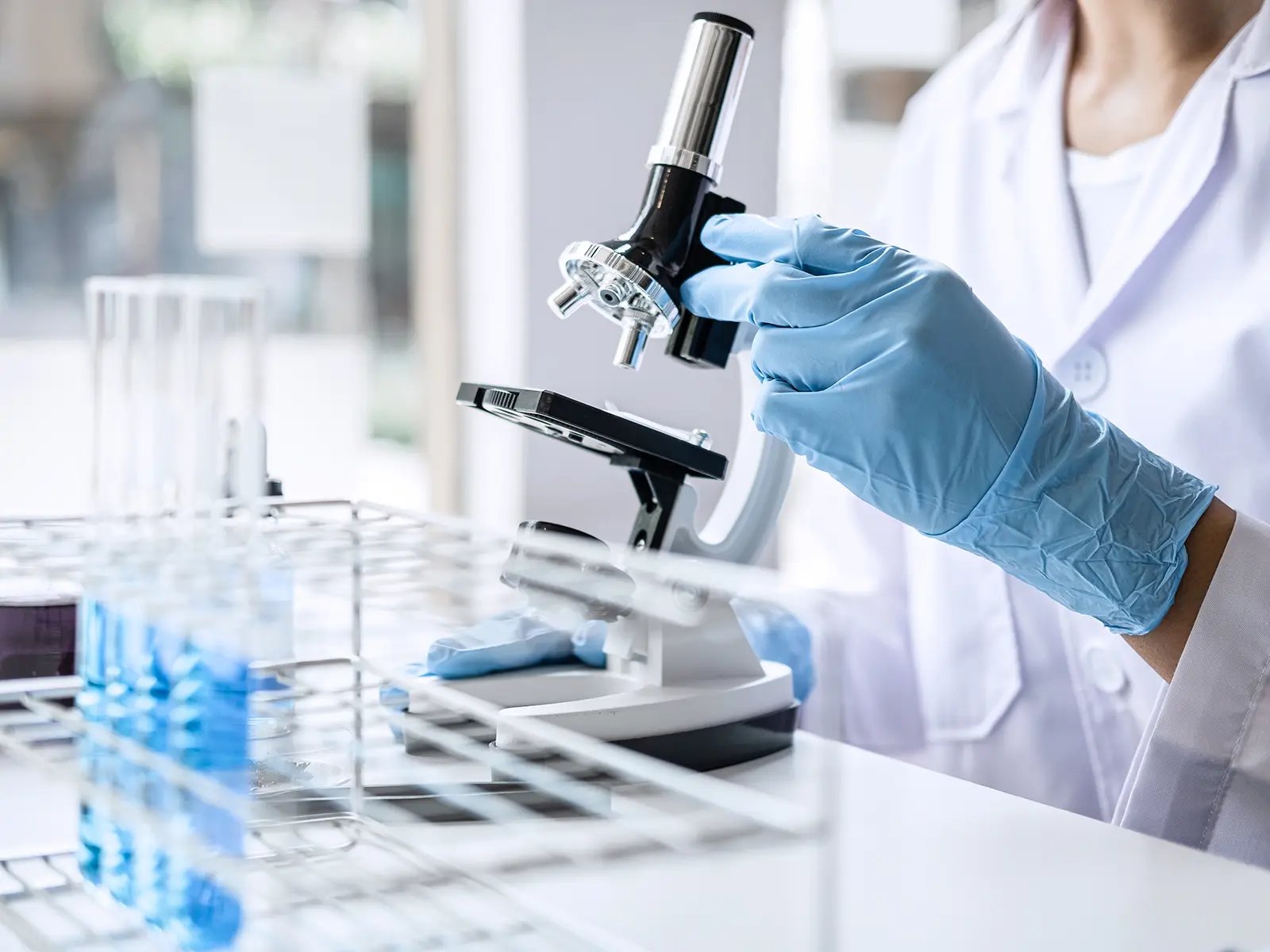 Profissional em laboratório clínico utilizando EPI e manipulando microscópio em bancada.