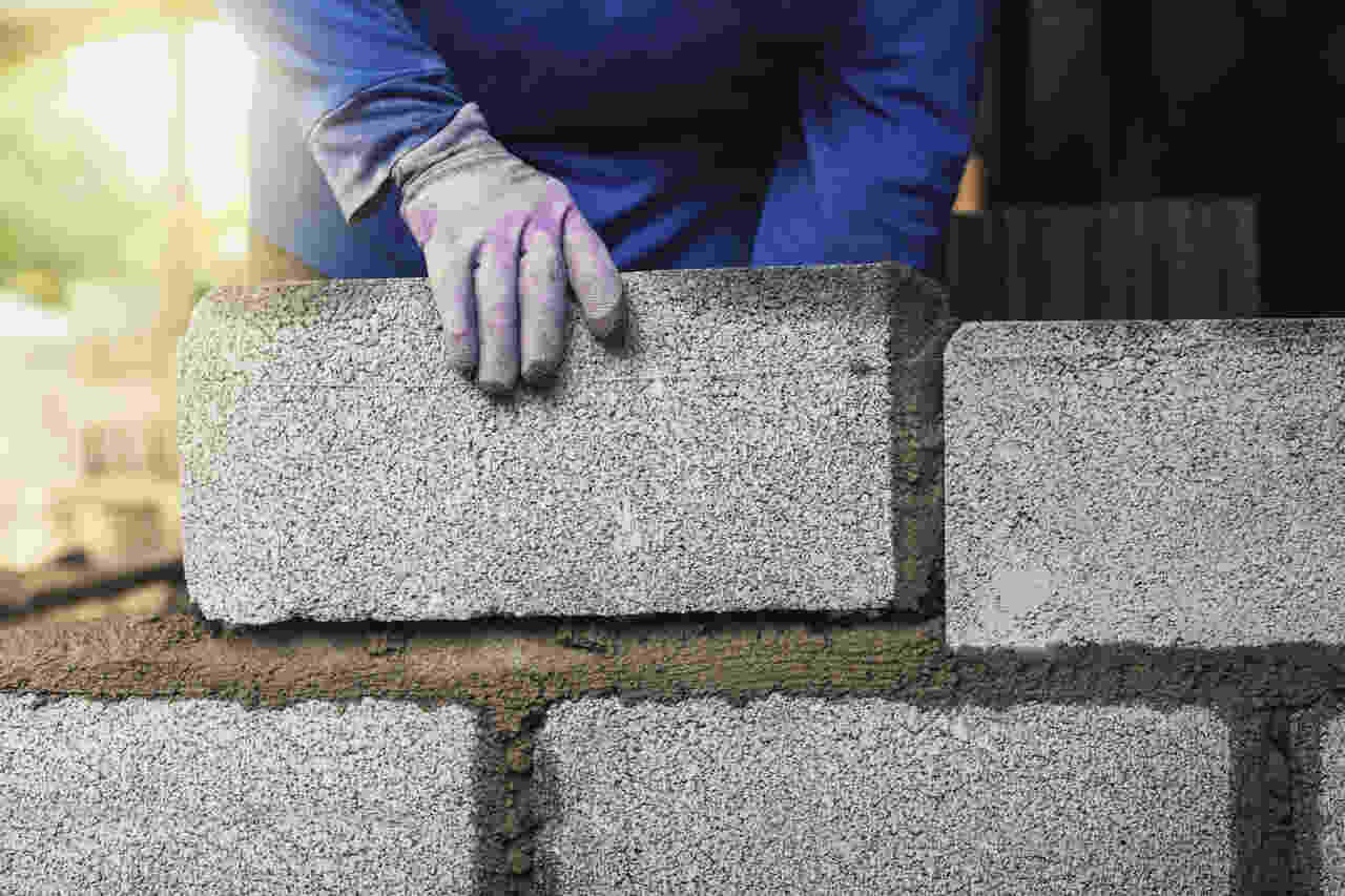 Pedreiro assentando blocos em obra urbana, utilizando EPI completo