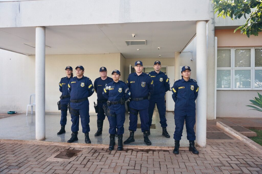 Equipe de guarda municipal com coletes azuis, em formação, à frente de prédio público
