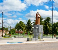 Praça e área urbana de Pirpirituba PB, com monumento central e arborização, em tarde ensolarada.