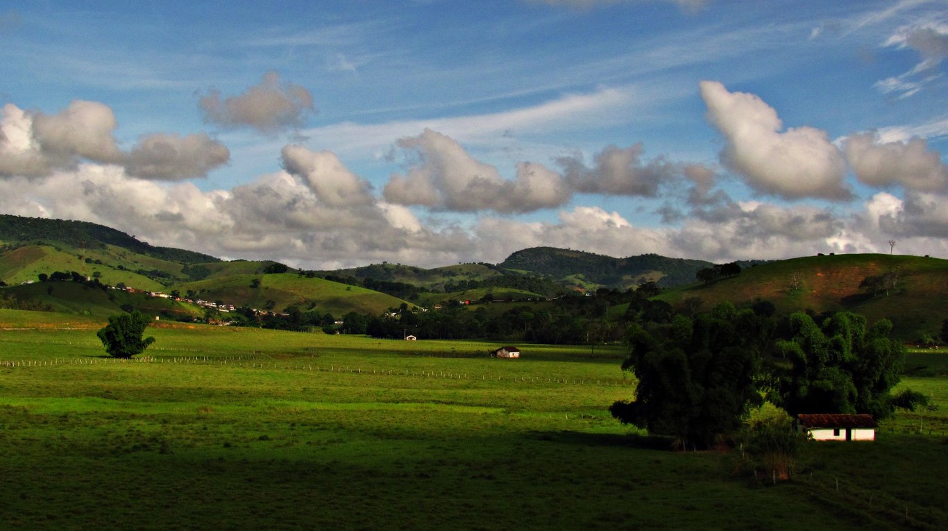 Paisagem urbana e rural da Zona da Mata mineira, com morros ao fundo, em orientação horizontal.