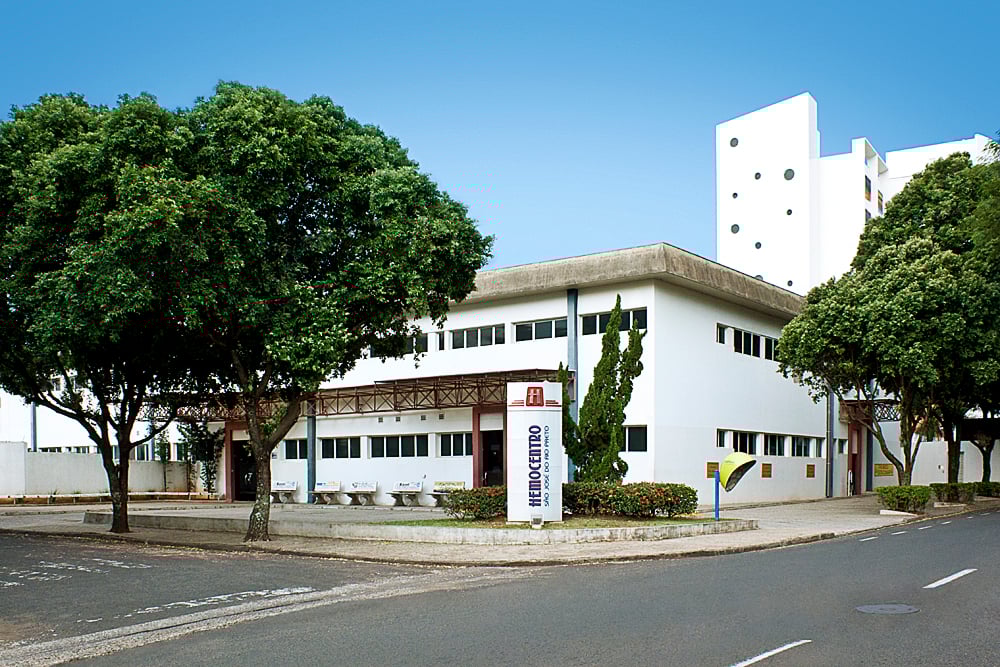 Foto da fachada de um hemocentro moderno em ambiente urbano. A imagem mostra árvores e movimento de pessoas ao redor do prédio.