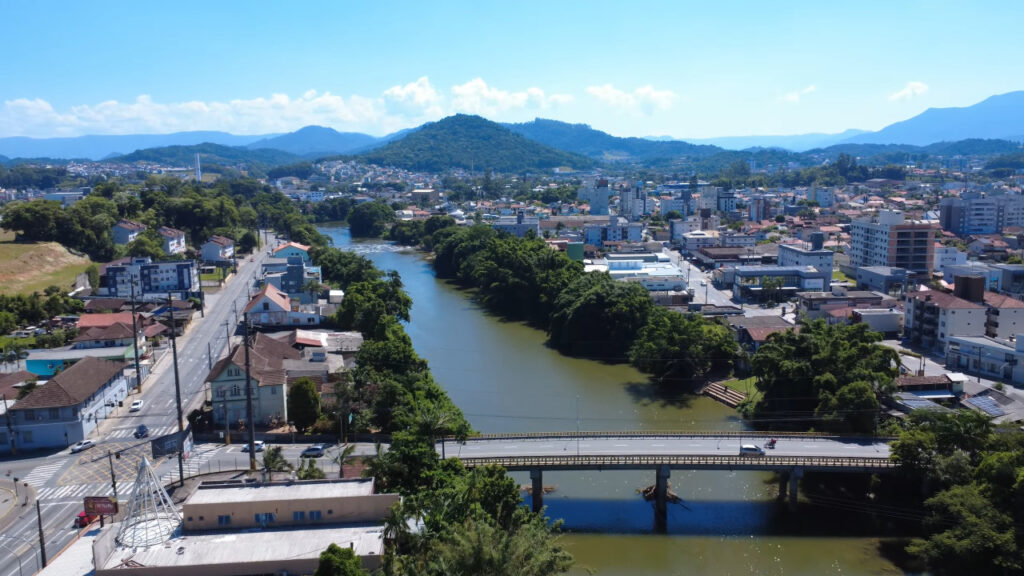 Vista aérea de Jaraguá do Sul com o rio ao centro e morros ao fundo, em dia claro
