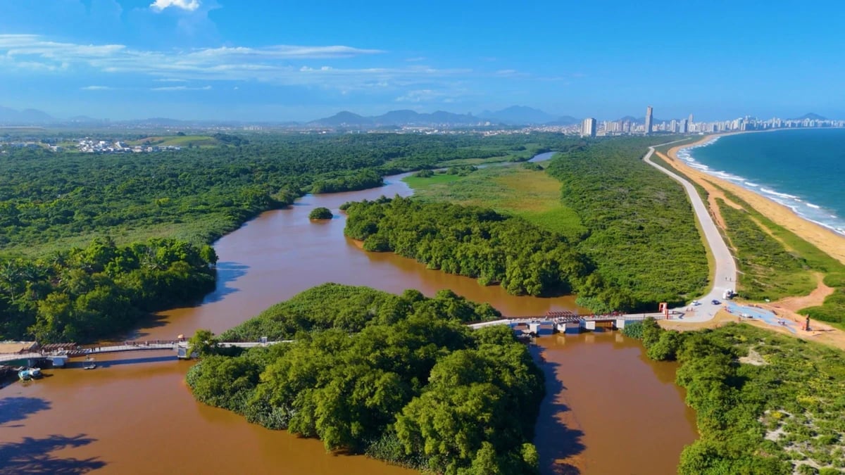 Vista aérea com áreas verdes, curso d’água e cidade ao fundo, na Grande Vitória (ES)