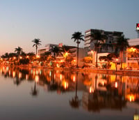 Vista urbana de Sete Lagoas ao entardecer, com lago em primeiro plano e iluminação urbana refletida na água.