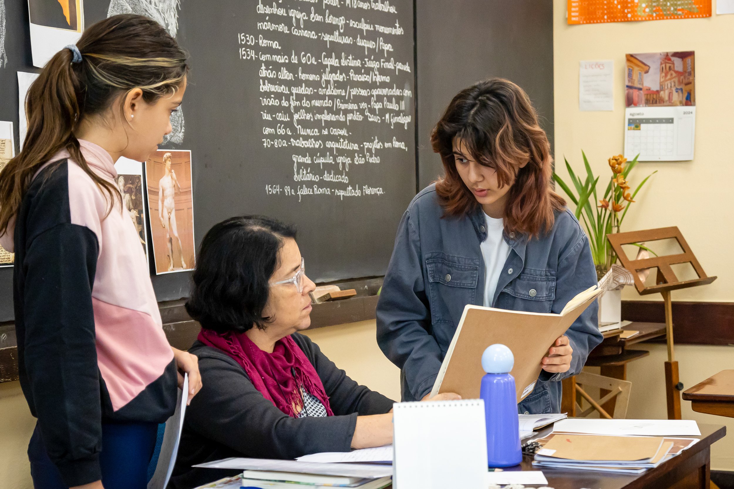 Sala de aula com professora e alunas, quadro e materiais didáticos