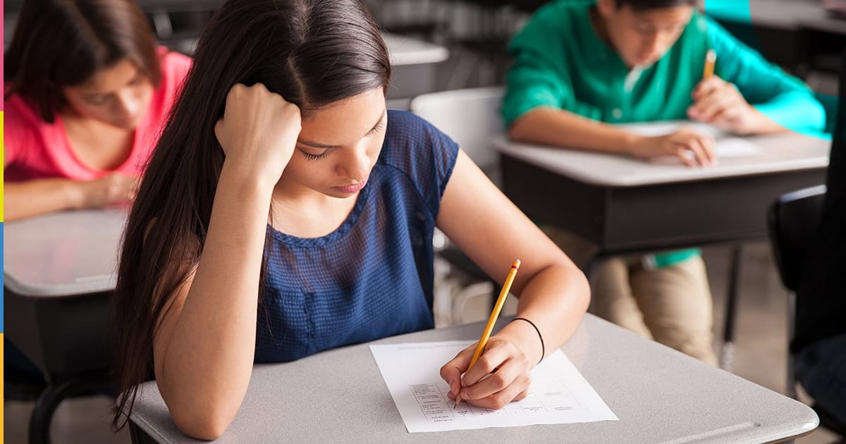 Sala de provas com carteiras alinhadas, cartão-resposta e caneta sobre a mesa, ambiente bem iluminado