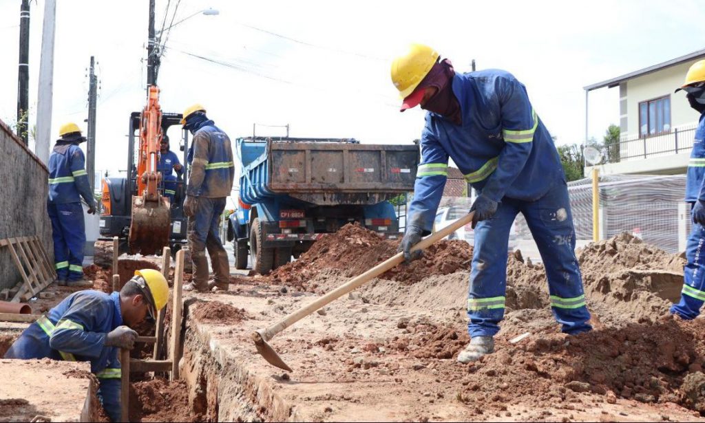 Profissionais de saneamento em atividade de campo, usando EPIs e coletores de dados, em área urbana
