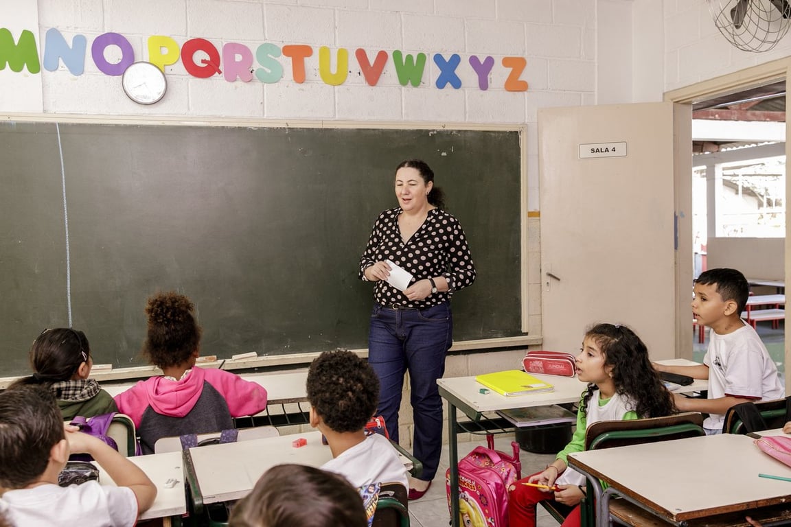 Professora em sala de aula do ensino fundamental escrevendo na lousa, enquanto alunos acompanham a aula. A imagem mostra um ambiente educacional com iluminação natural.