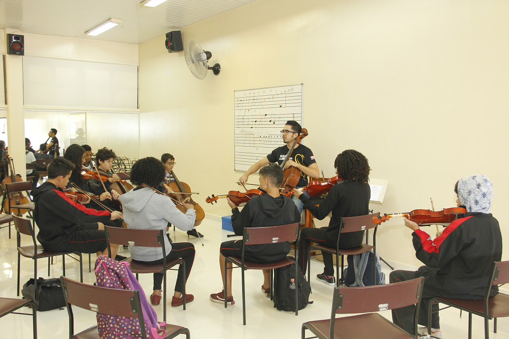 Sala de aula de música com alunos tocando violinos e professor orientando, com instrumentos de percussão ao fundo