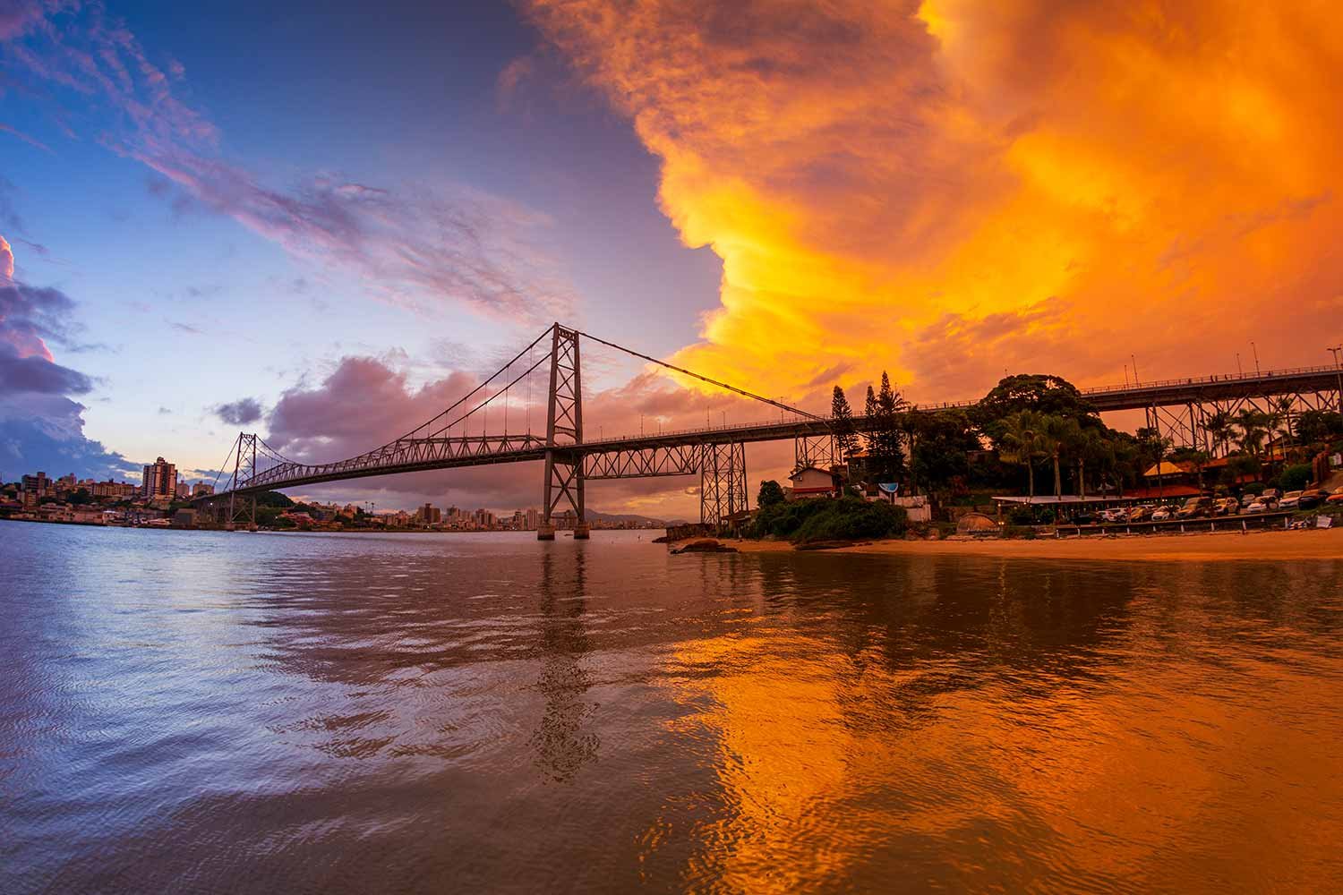 Vista aérea de Florianópolis ao entardecer, com a Ponte Hercílio Luz em destaque e céu alaranjado refletindo no mar.