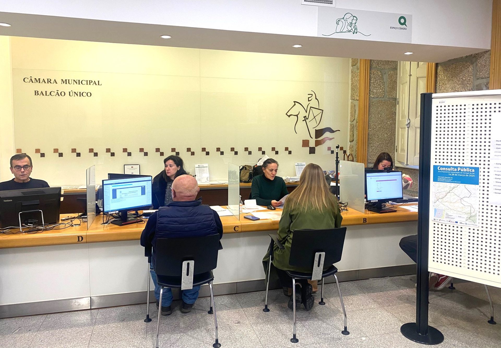 Ambiente de atendimento ao público em repartição municipal, com balcão, computadores e documentos