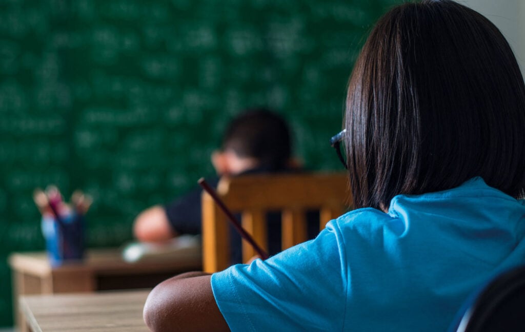 Sala de aula em escola pública, quadro ao fundo e estudantes em foco desfocado