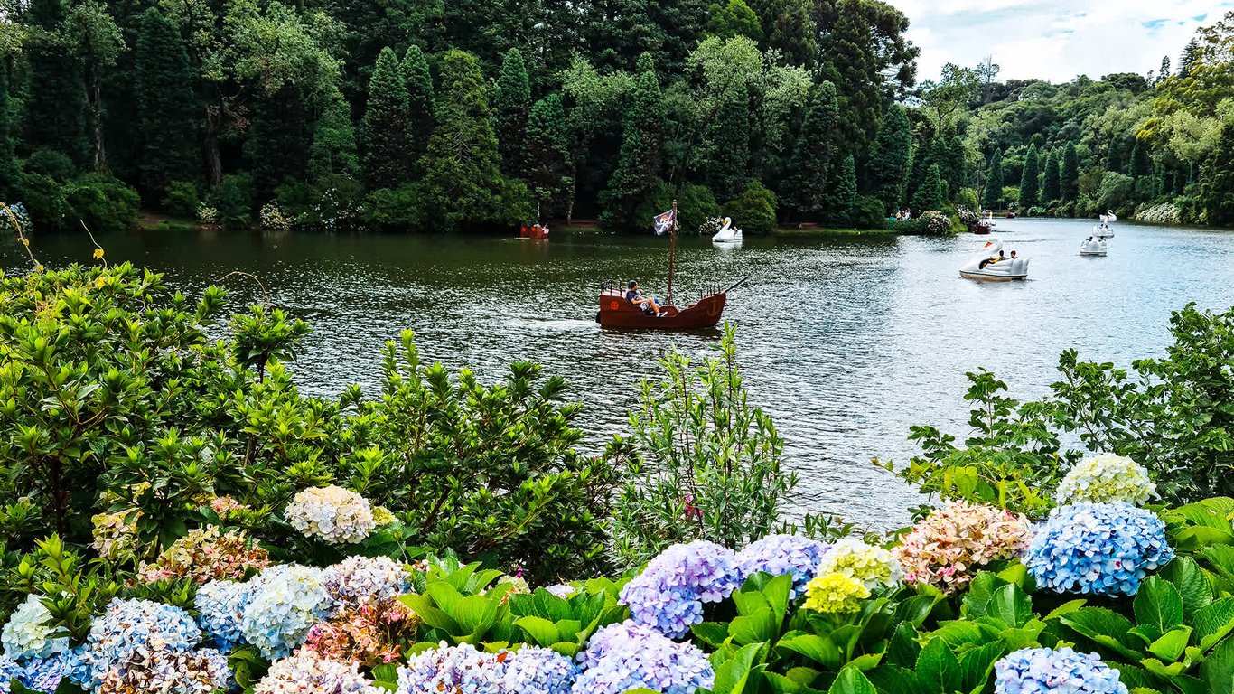 Cenário de Gramado RS, com arquitetura de influência europeia e paisagismo florido à beira de lago, em dia ensolarado