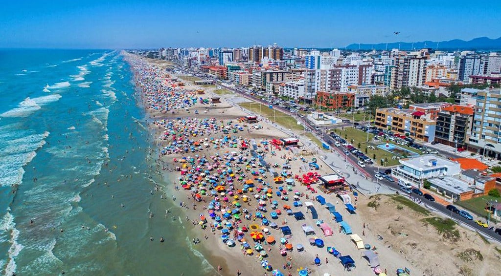 Vista do litoral de Xangri-Lá (RS), com faixa de areia e prédios ao fundo, em dia ensolarado.