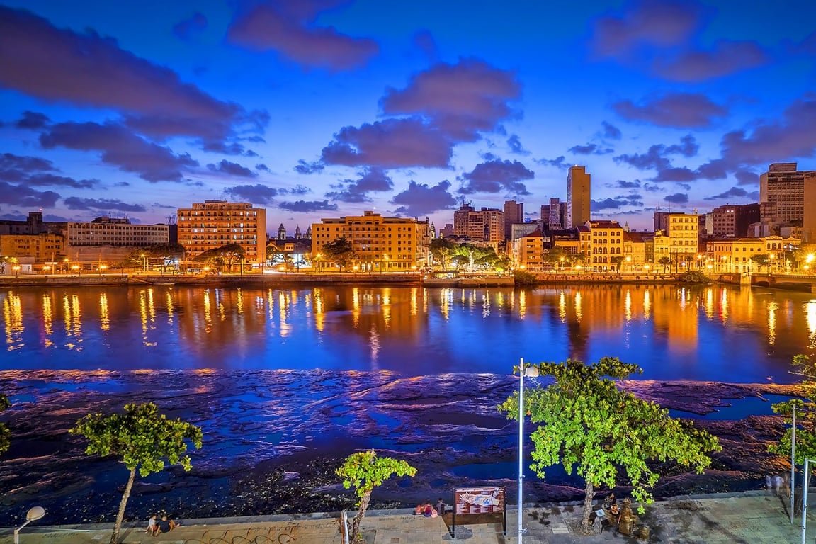 Vista aérea de Recife ao entardecer, com o Rio Capibaribe e prédios modernos ao fundo.
