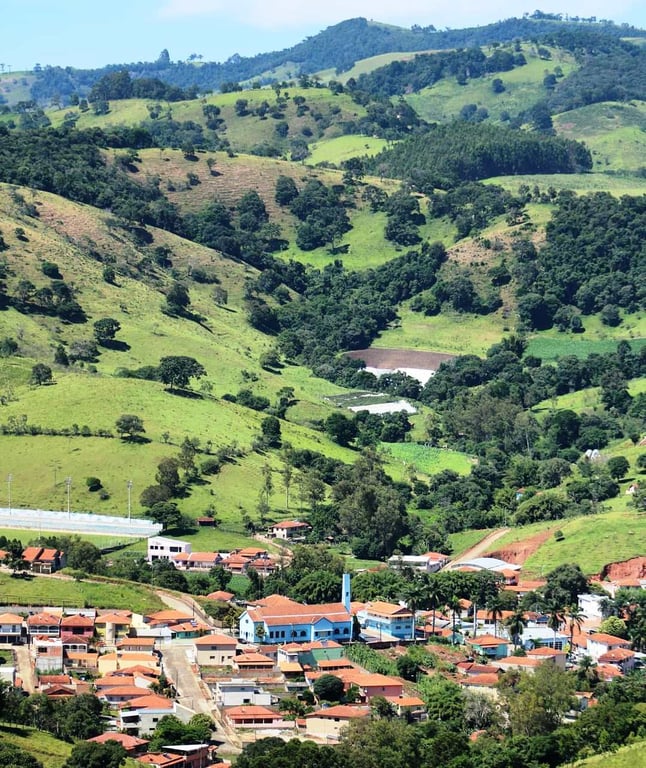 Vista urbana pitoresca de uma vila rural cercada por colinas verdes. Há várias casas com telhados vermelhos e uma igreja azul, típica de regiões cafeeiras de Minas Gerais, transmitindo uma sensação de tranquilidade e paisagem interiorana.