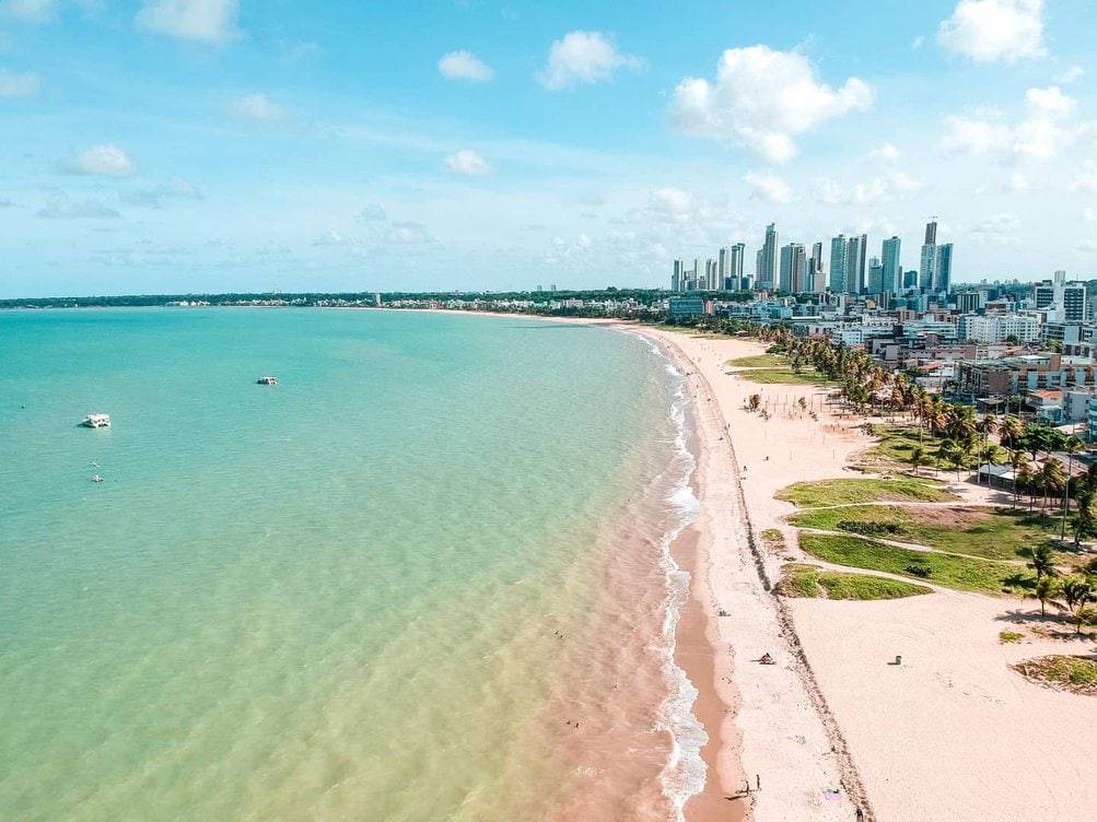 Vista aérea da orla urbana na Paraíba, com faixa de praia e edifícios sob céu azul.