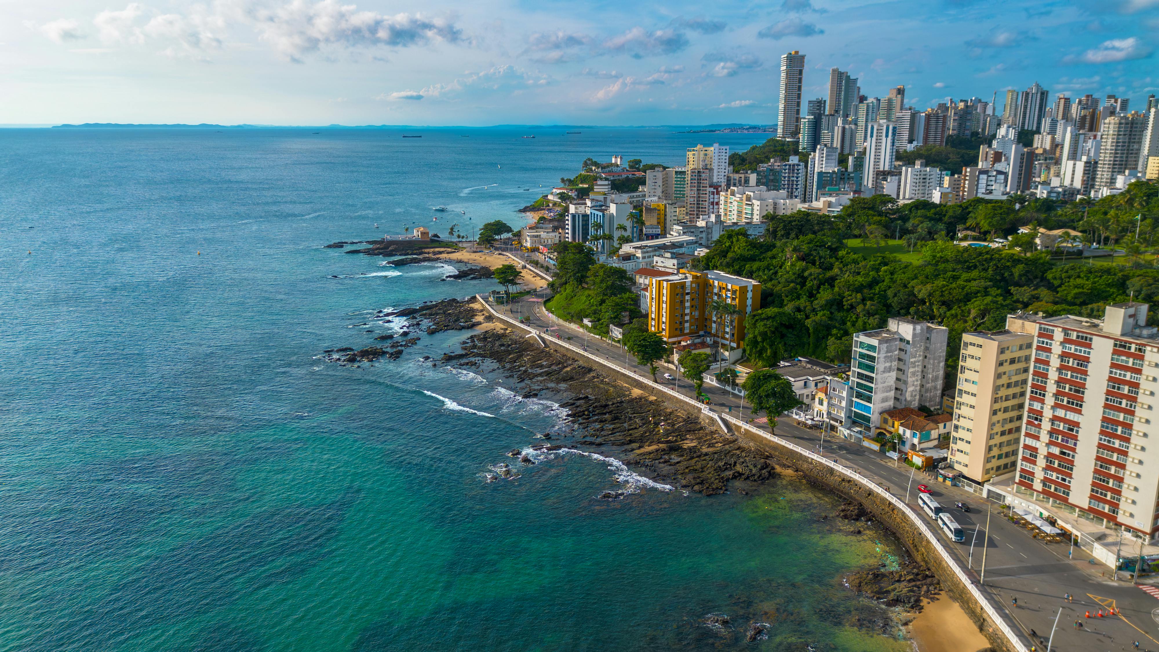 Vista aérea de Salvador, com orla, avenidas e céu azul; trânsito moderado na via à beira-mar