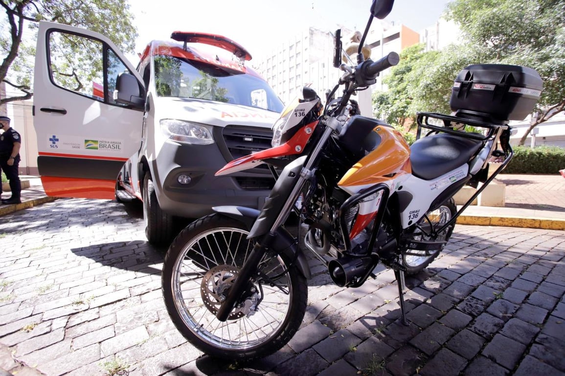 Motolância do SAMU em operação urbana, com profissional ao lado da moto e viatura de apoio ao fundo