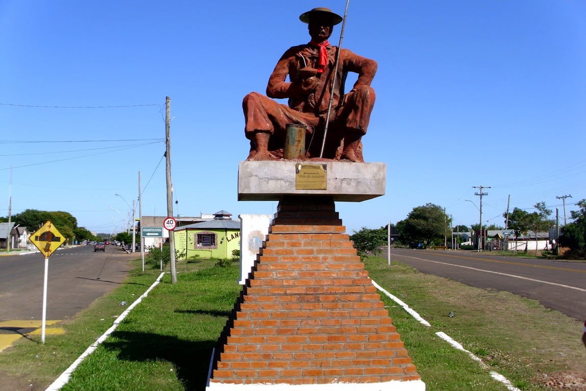 Cenário urbano-ribeirinho na Fronteira Oeste do RS, com monumento e avenida arborizada sob céu aberto