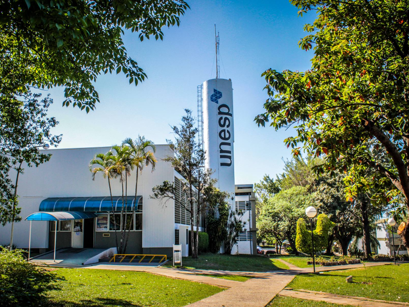Entrada do campus da Unesp em Marília, com letreiro da universidade em destaque e vegetação ao redor em um dia claro.
