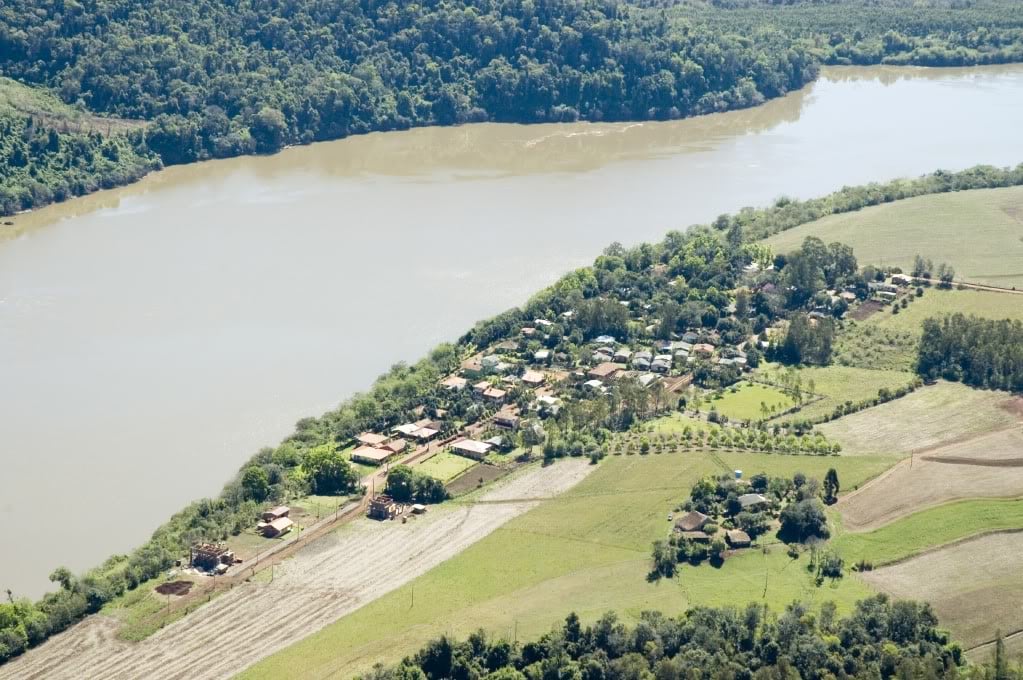 Vista aérea de uma pequena cidade no interior do Rio Grande do Sul, com ruas e construções baixas cercadas pela natureza, em dia claro.