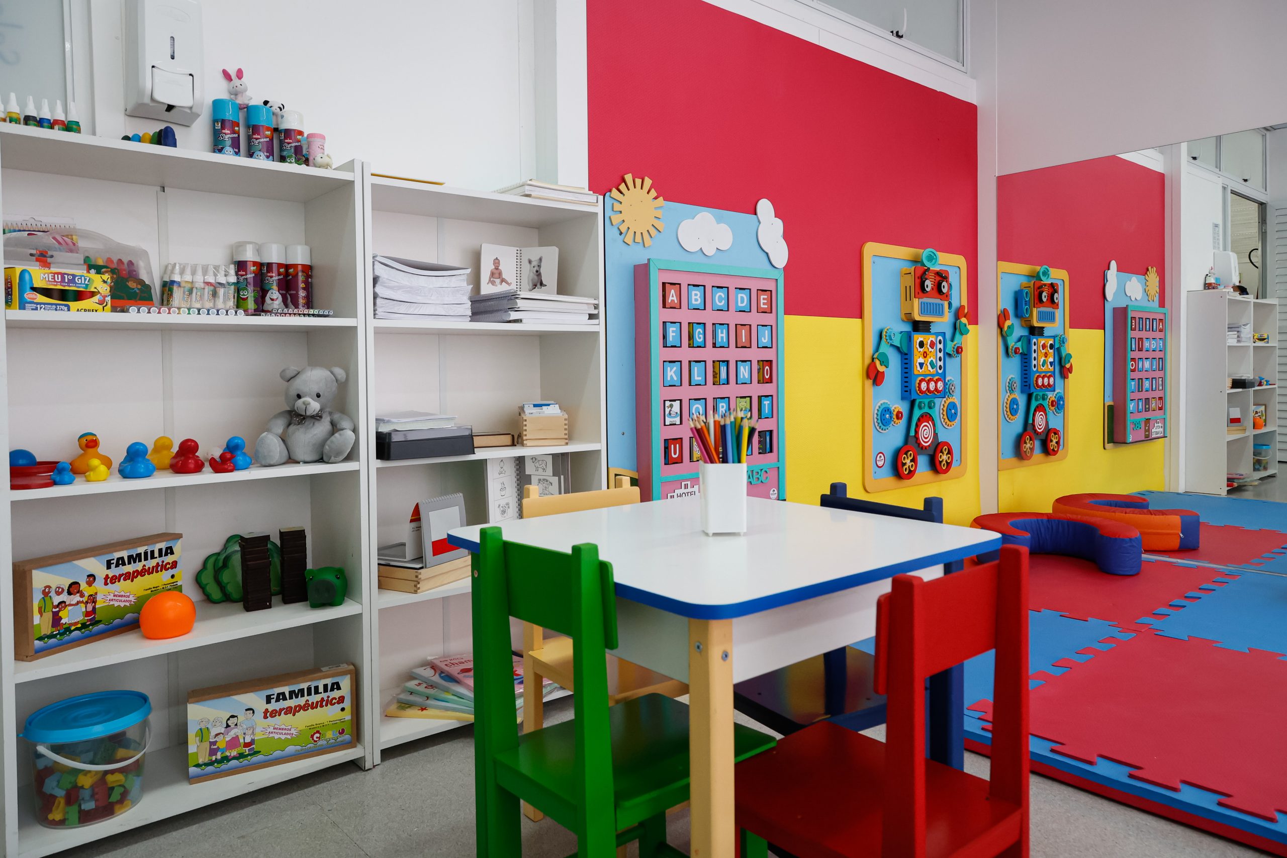 Consultório com mesa, cadeiras coloridas e prateleiras com materiais lúdicos, remetendo a atendimentos de reabilitação