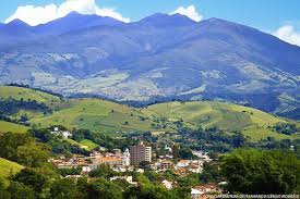 Vista aérea de uma cidade mineira cercada por montanhas e vegetação, com clima de cidade pequena. Mostra ruas e casario característicos da região.