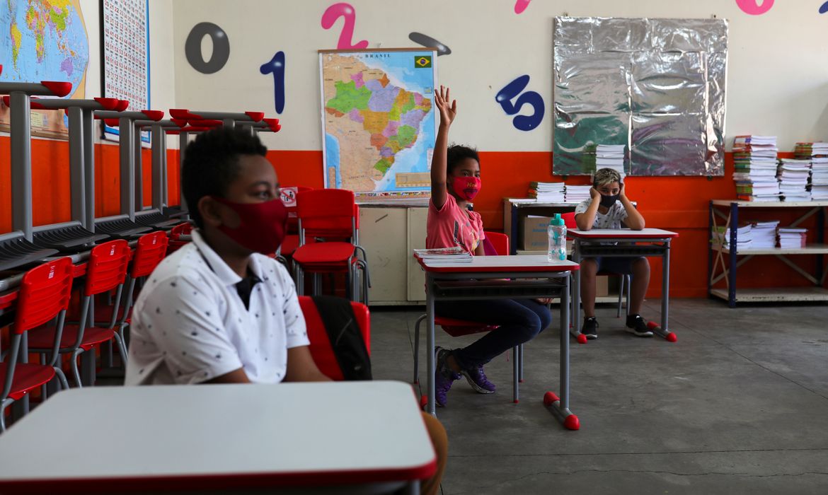 Sala de aula com estudantes adultos em ambiente escolar moderno; um aluno levanta a mão para falar