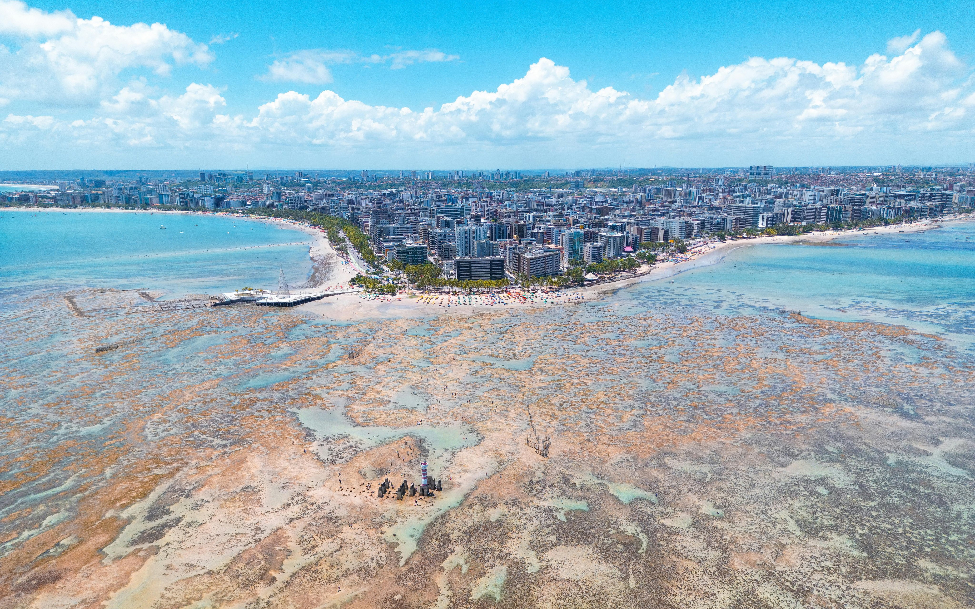 Vista aérea de Maceió, com orla, mar azul e prédios ao fundo, ao pôr do sol