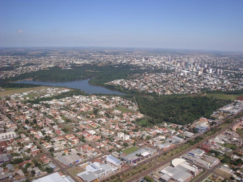 Vista aérea urbana no Oeste do Paraná, com quarteirões, áreas verdes e rio ao entardecer.