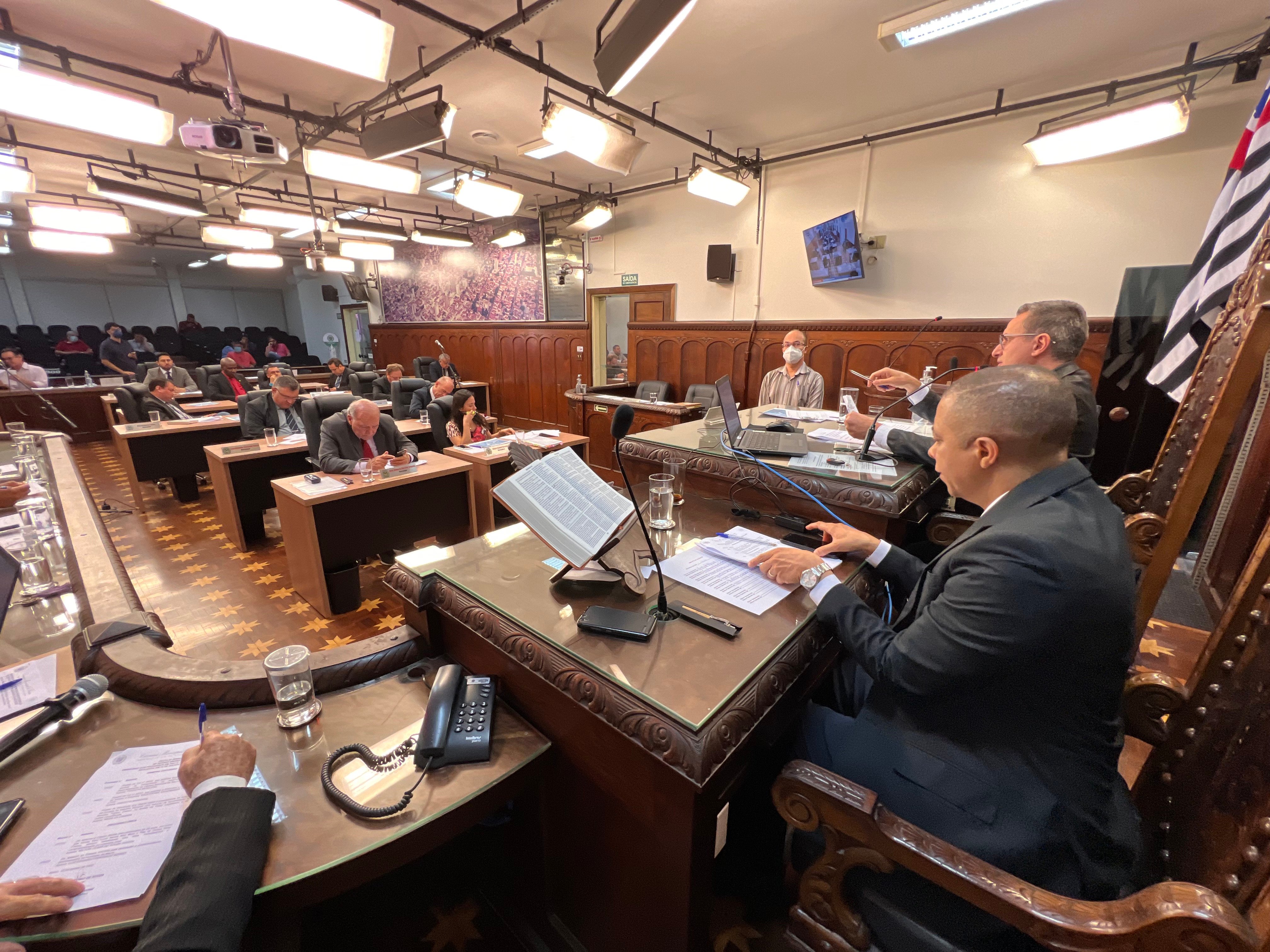 Interior de um plenário de câmara municipal, com vereadores em sessão; microfones e mobiliário ao fundo.