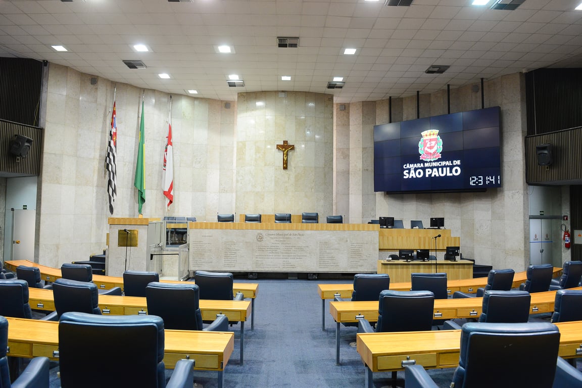 Interior de plenário de uma câmara municipal brasileira, com bandeiras e brasão ao fundo
