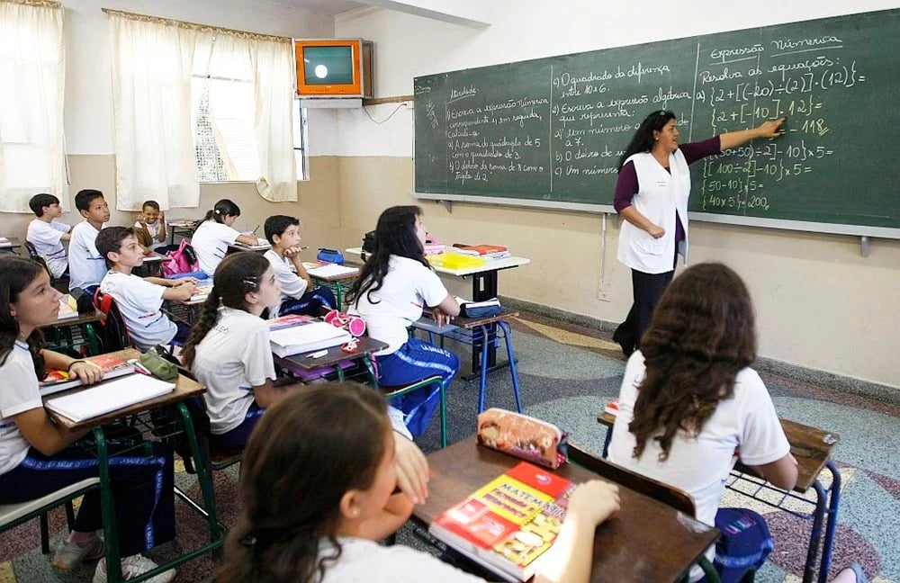 Sala de aula do ensino fundamental com professora escrevendo no quadro e alunos atentamente acompanhando a explicação