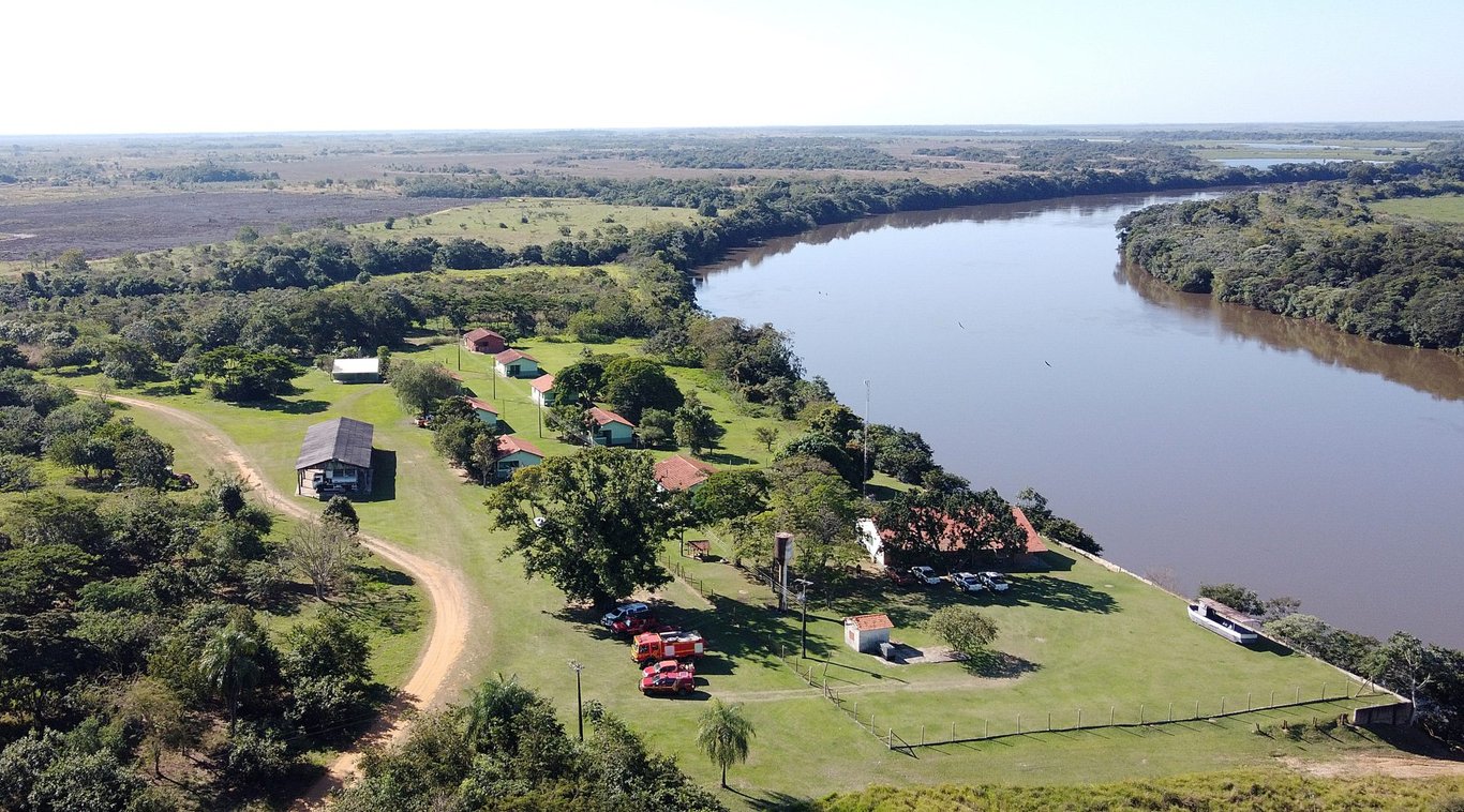 Paisagem pantaneira com áreas alagadas e vegetação típica; ao fundo, estruturas e veículos na margem