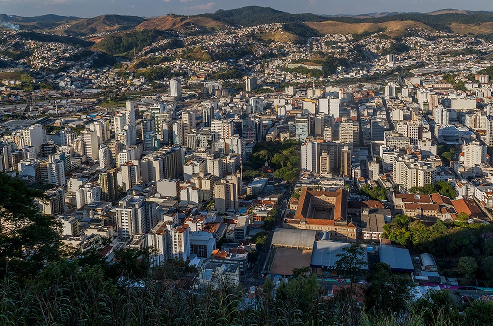 Vista aérea de cidade mineira entre morros, com área urbana e vegetação ao redor, em fim de tarde