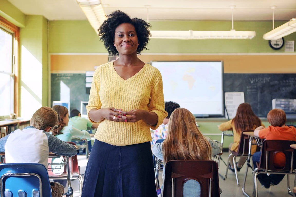 Sala de aula na rede pública brasileira, com professora à frente e alunos de costas, em ambiente iluminado