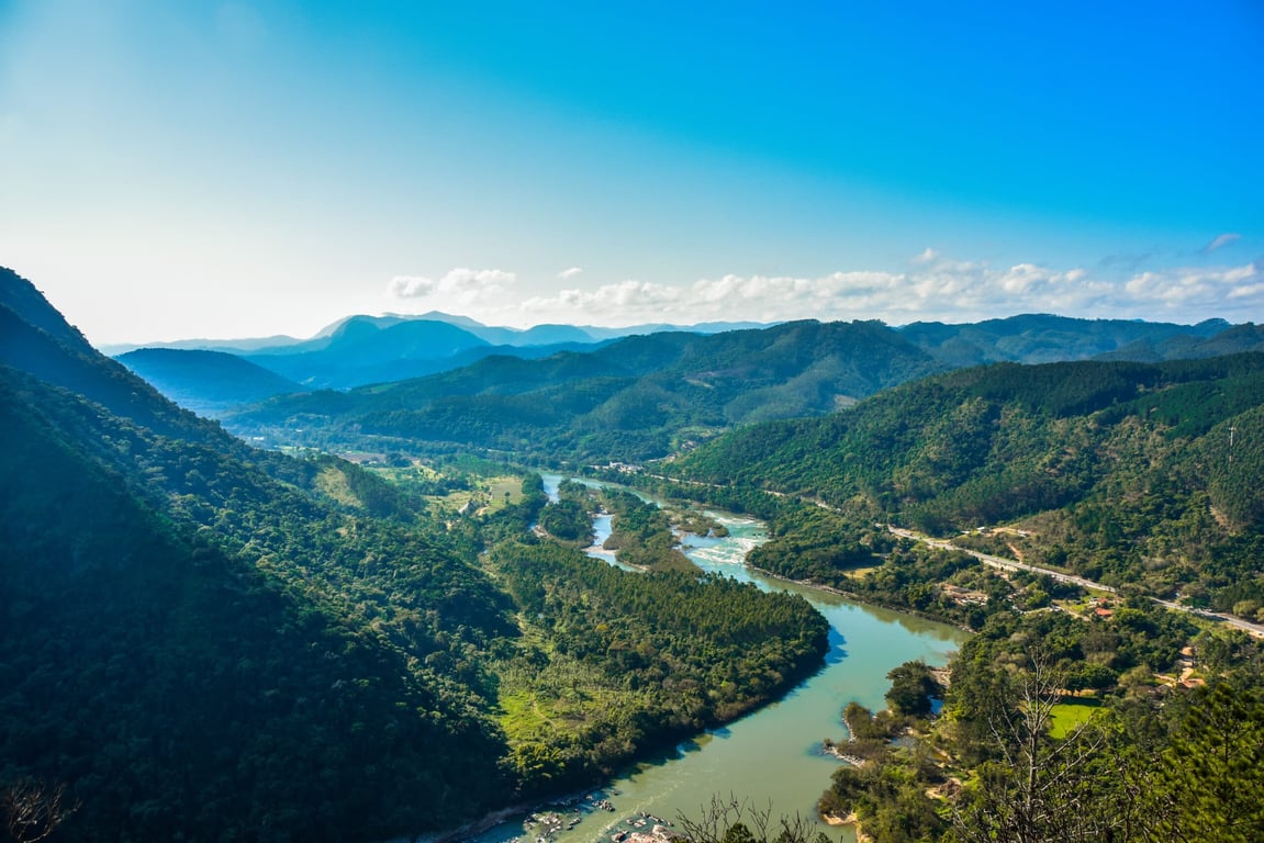 Paisagem do Vale do Itajaí com um rio serpenteando entre montanhas verdes. A cena é clara e bem iluminada, simbolizando a natureza da região de Apiúna SC.