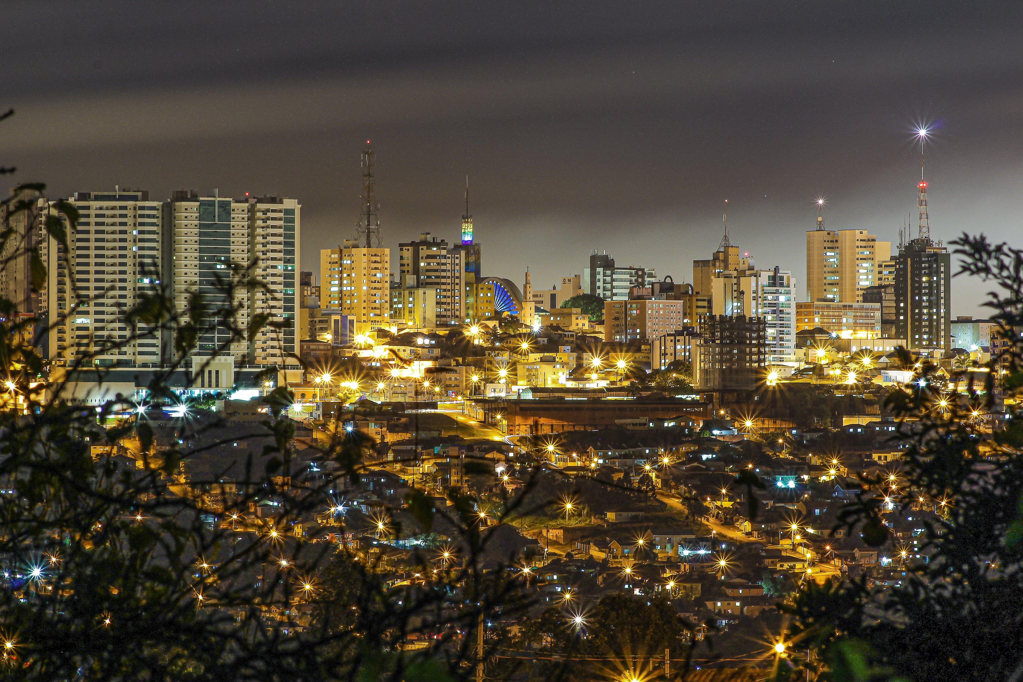 Vista urbana do interior do Paraná, à noite, com prédios baixos e vegetação iluminada. Foto ilustrativa.