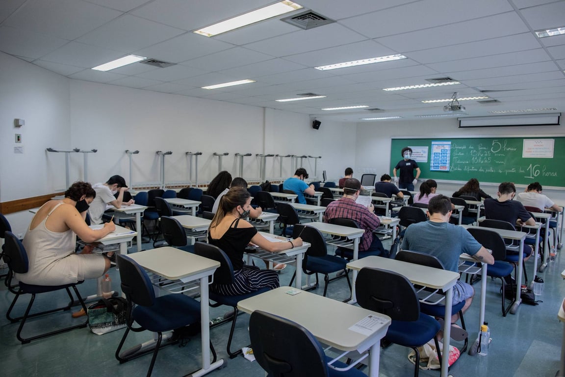 Sala de provas com candidatos realizando exame em carteiras, ambiente educacional iluminado