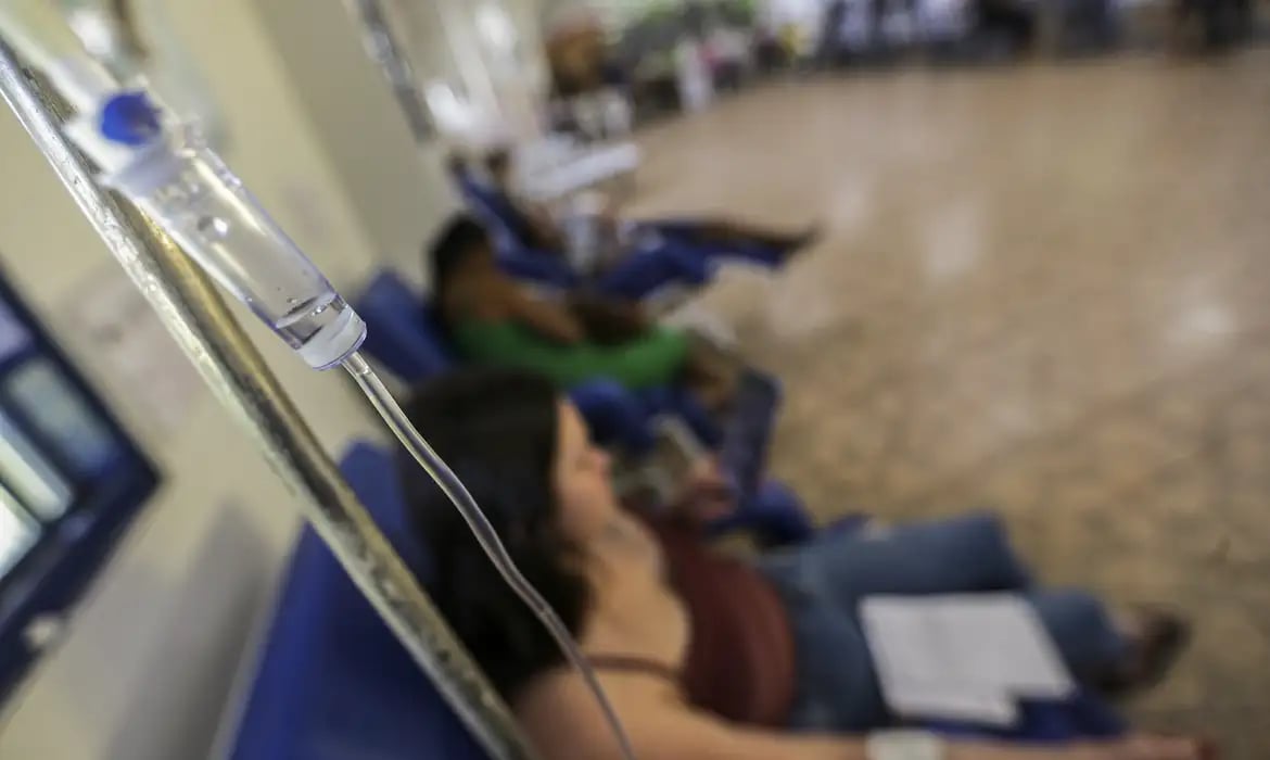 Recepção em unidade pública de saúde, com pacientes aguardando atendimento e profissional uniformizado ao fundo
