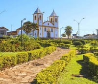 Paisagem urbana de Queluzito MG com igreja colonial e morros ao fundo sob céu azul.