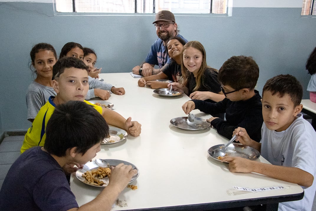 Refeitório escolar com bandejas de merenda saudável; crianças desfocadas ao fundo e adulto supervisionando