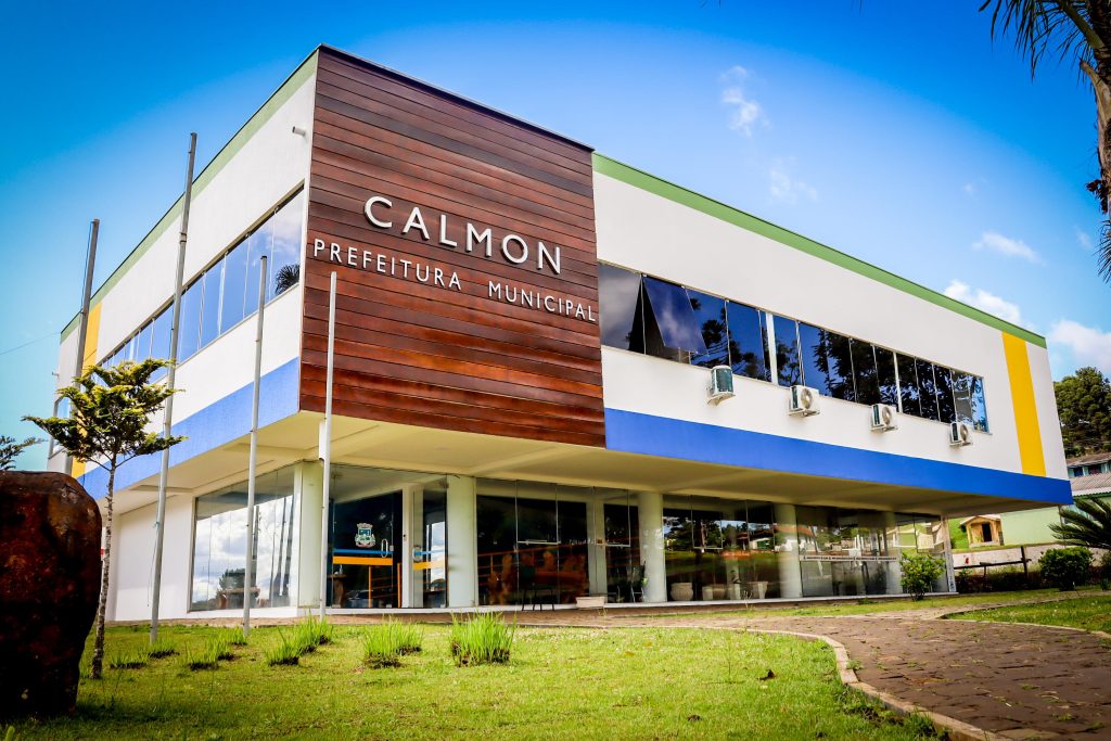 Fachada da Prefeitura de Calmon, com bandeiras e arquitetura moderna, em dia ensolarado