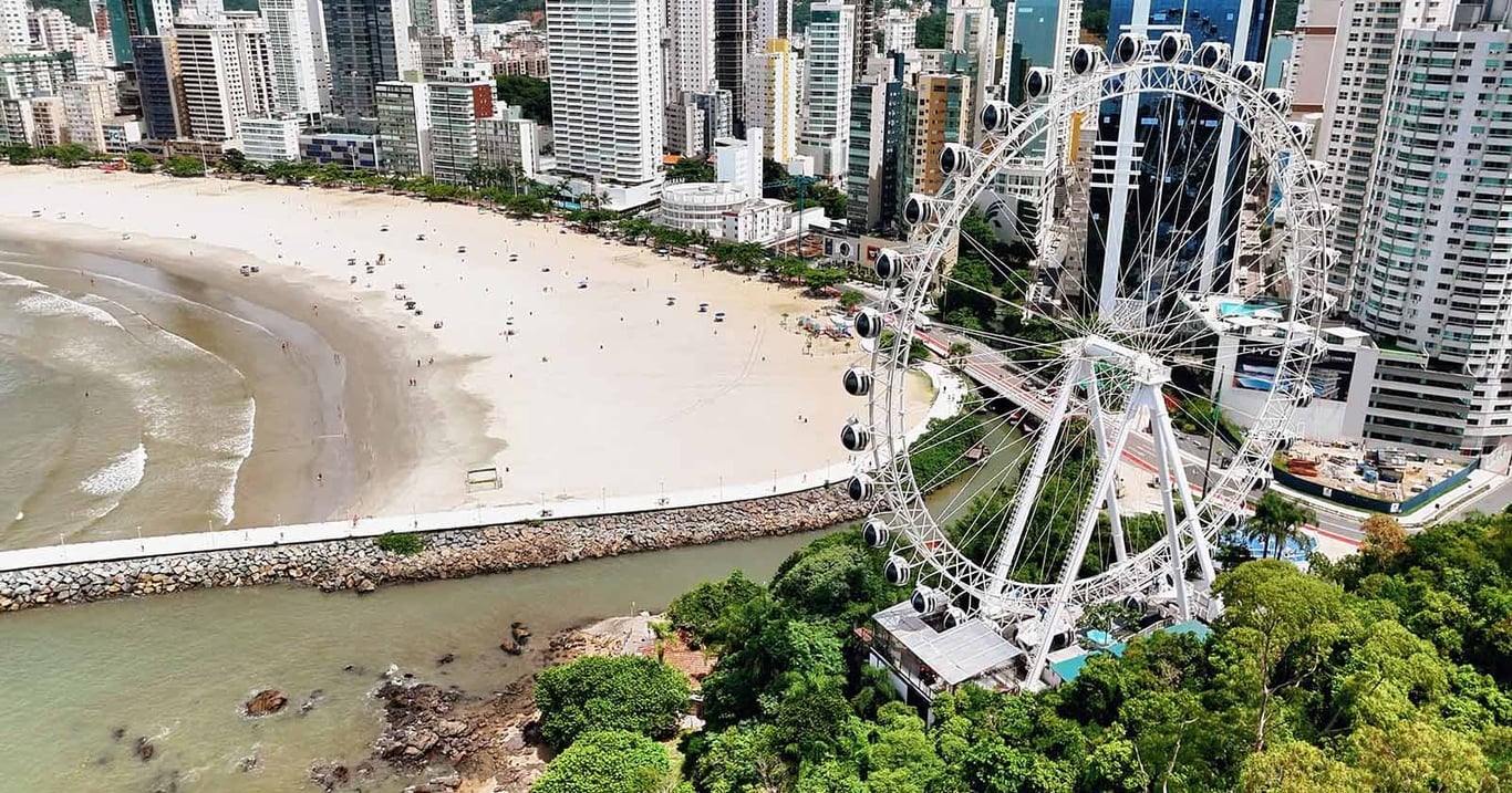 Vista aérea de Camboriú com a orla e arranha-céus ao fundo, mostrando a roda-gigante e a praia movimentada
